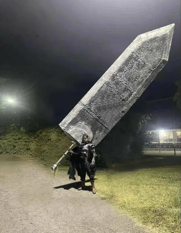 A person in dark, possibly fantasy-themed clothing stands outdoors at night on a paved path, carrying an enormous, crudely…