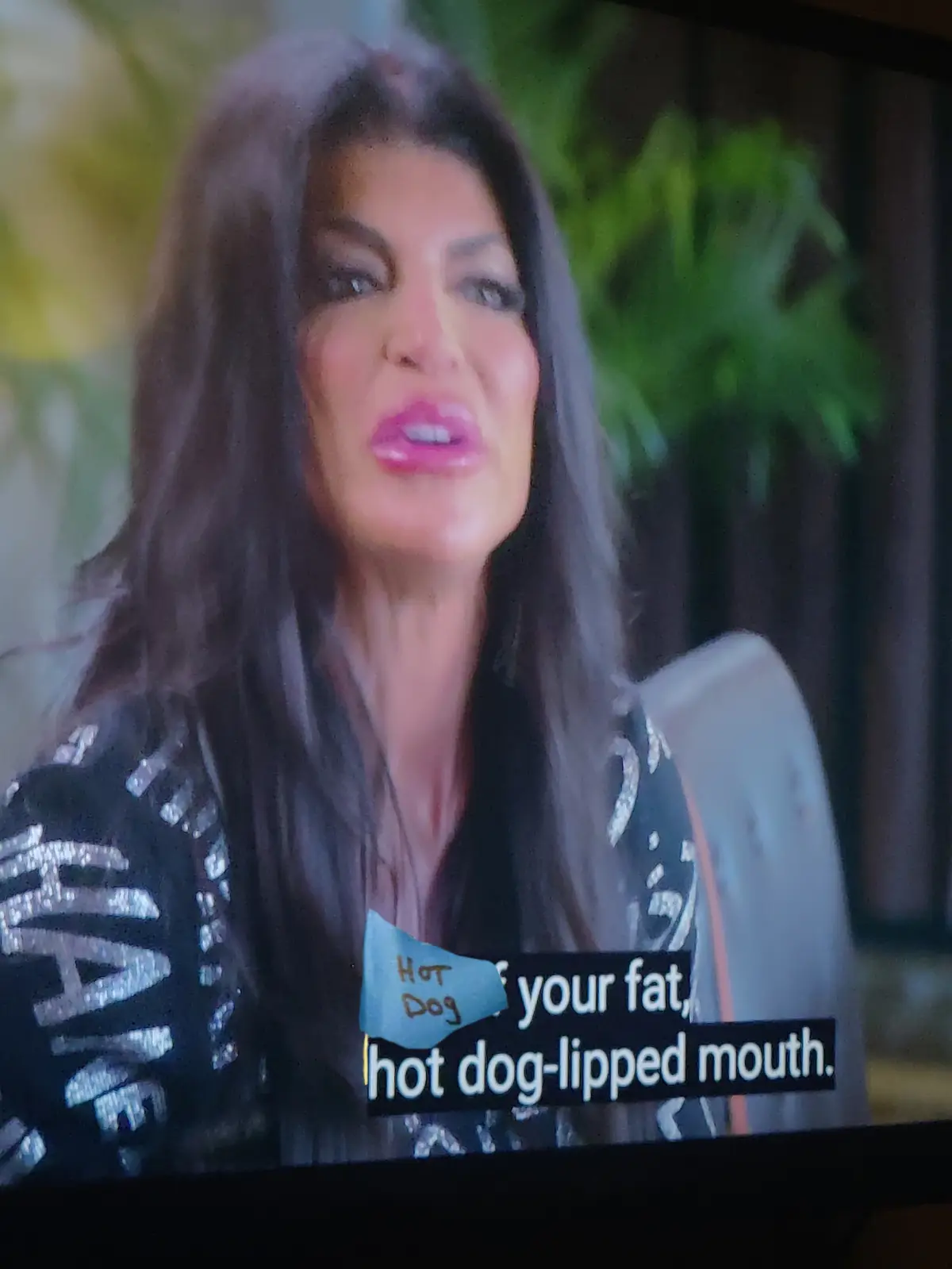 A close-up of a woman with long dark hair and pink lipstick, wearing a black jacket with silver lettering, appears to be…