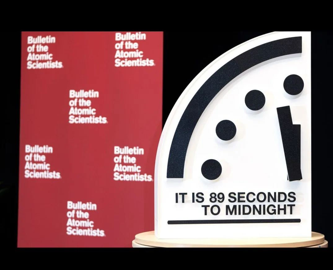 A white, semi-circular Doomsday Clock display with black hour and minute hands, showing the time as 89 seconds to midnight…