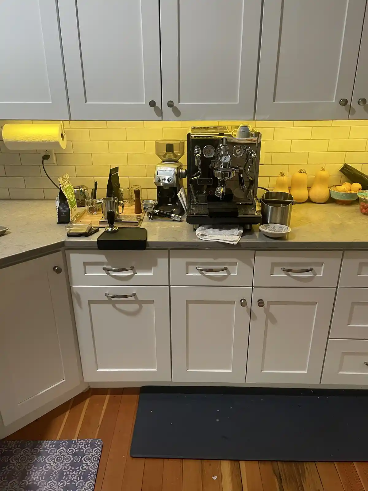A kitchen counter with white cabinets and a yellow tiled backsplash, featuring a professional espresso machine, grinder, and…