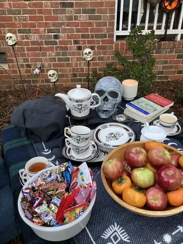 An outdoor Halloween tea party setup features a table with a skull-themed teapot and cups, a large decorative skull, bowls…