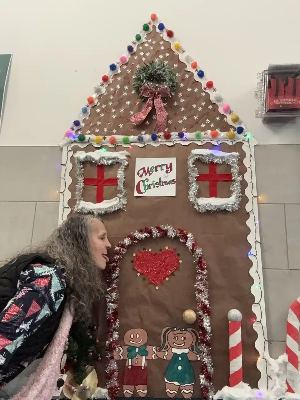 A woman with long gray hair sticks her tongue out playfully while standing beside a large, decorated gingerbread house. The…