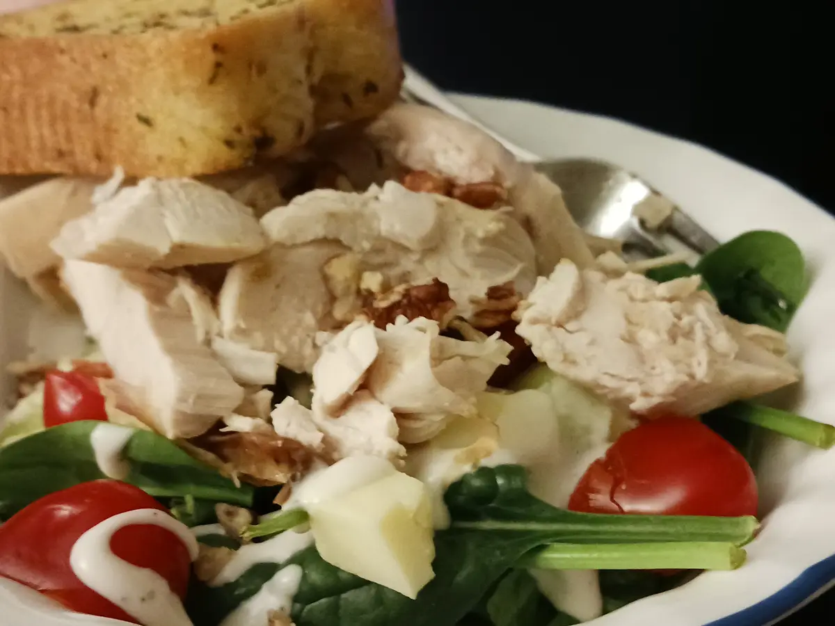 A close-up photo of a plated salad with spinach, cherry tomatoes, shredded chicken, croutons, and a creamy dressing, with a…