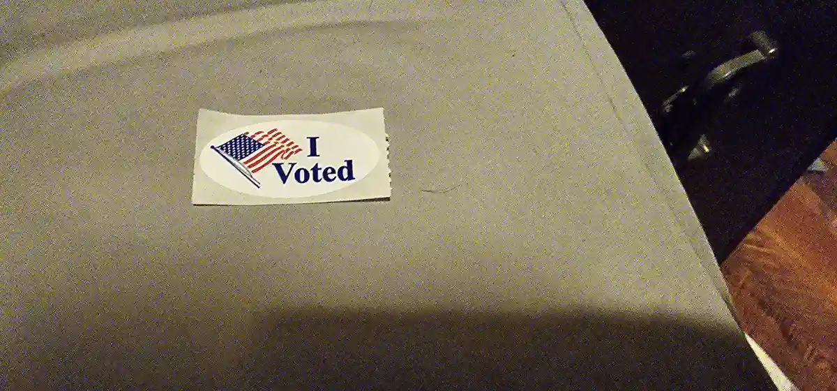 A close-up photo shows a white oval 'I Voted' sticker with an American flag graphic, lying on a plain gray fabric surface…