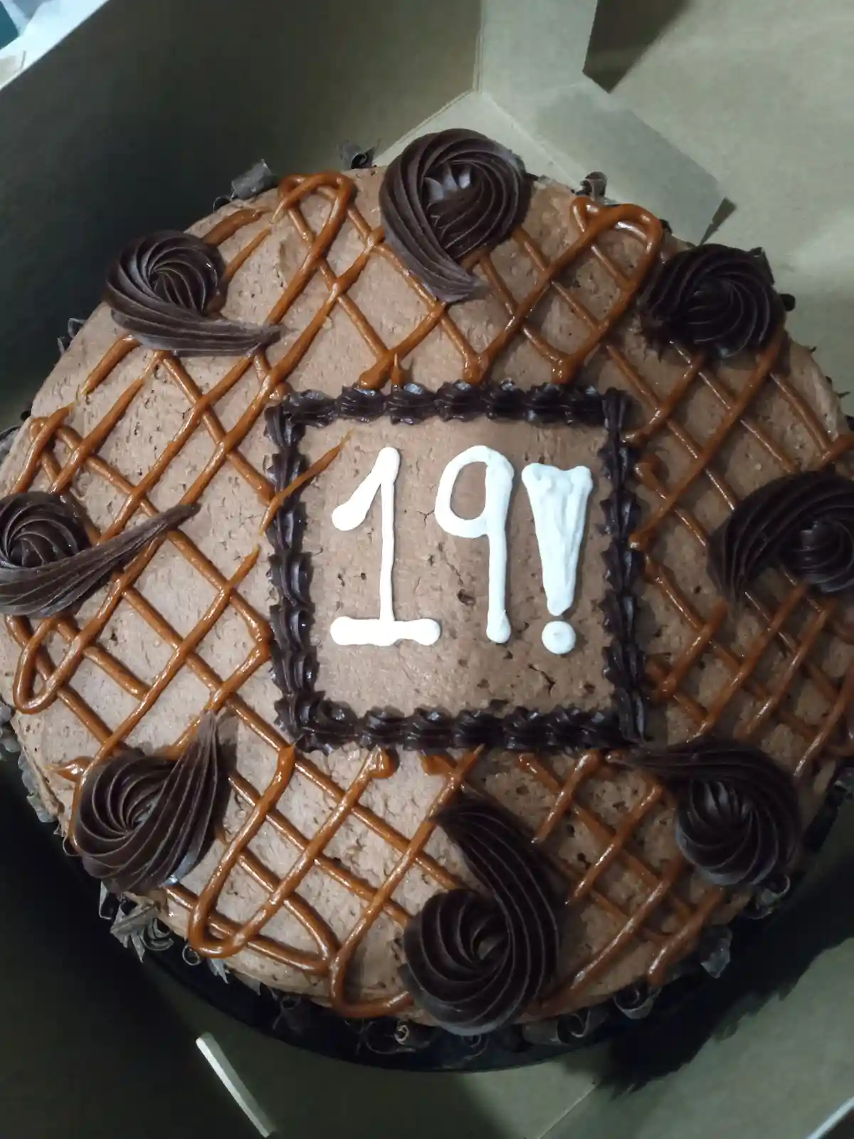 A round chocolate cake with caramel drizzle and chocolate swirls is decorated with the number '19!' written in white icing…