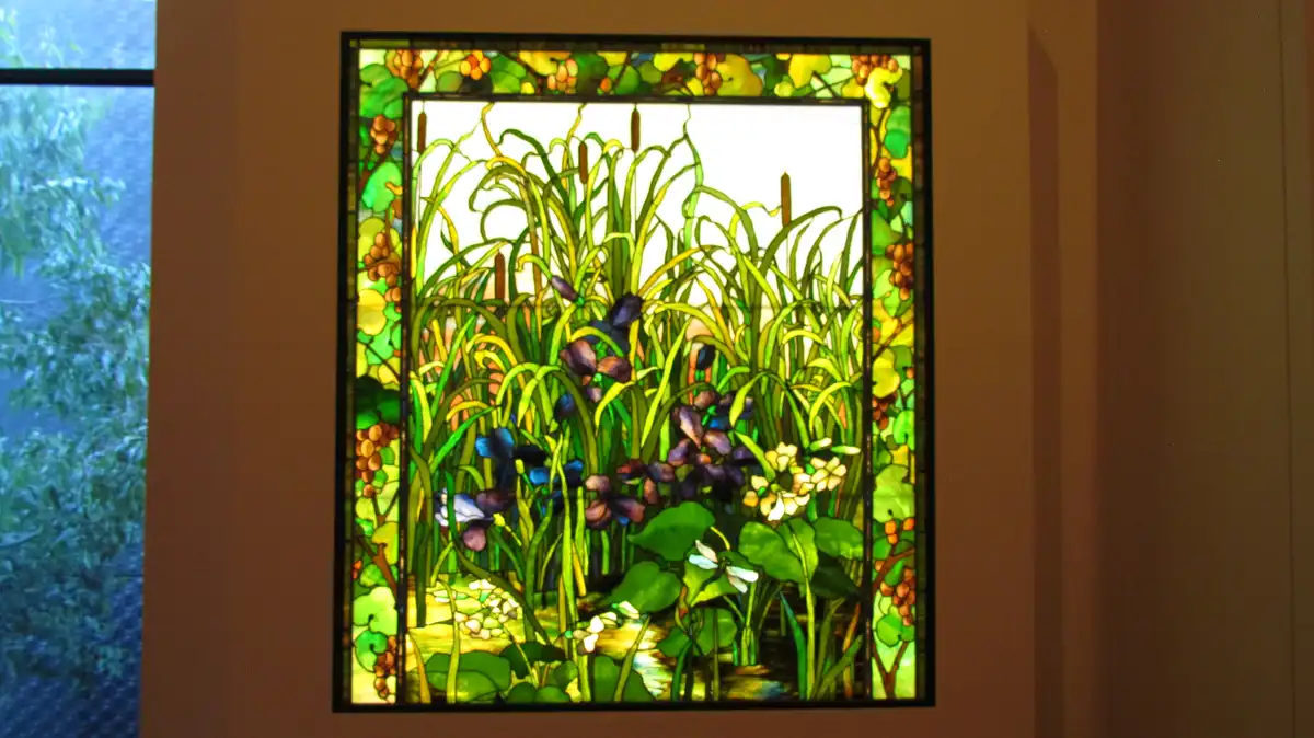 a stained glass window depicts a vibrant garden scene with tall green reeds, purple and blue flowers, white blossoms, and a…