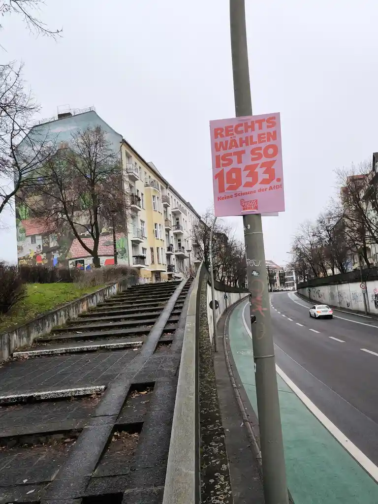 A pink political poster is affixed to a street pole in an urban setting, featuring bold red text that reads 'RECHTS WÄHLEN…