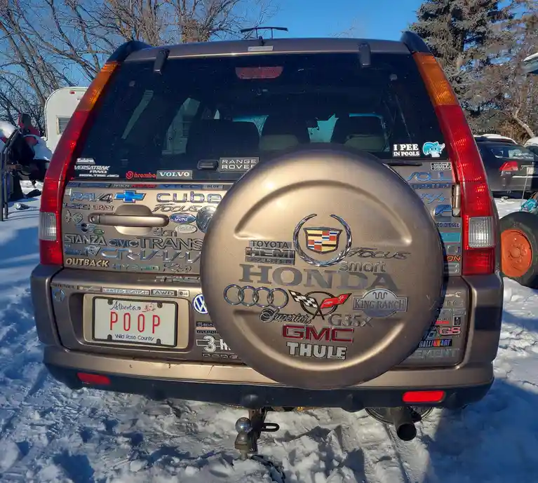 The rear of a brown SUV covered in numerous car brand stickers, including Toyota, Honda, GMC, and Cadillac, with a spare…