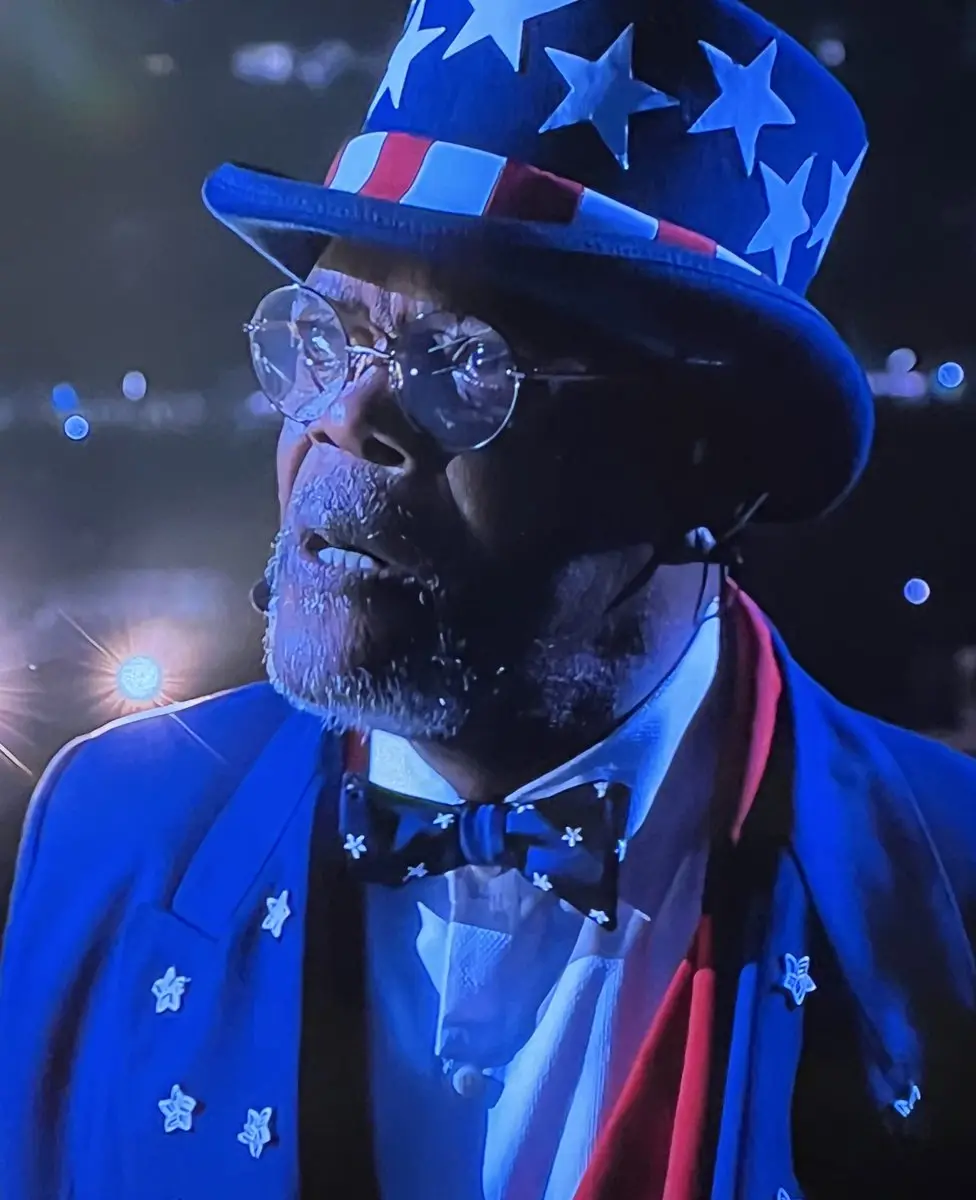 a man wearing a patriotic costume with a star-spangled top hat, round glasses, and a blue suit adorned with stars, looks…