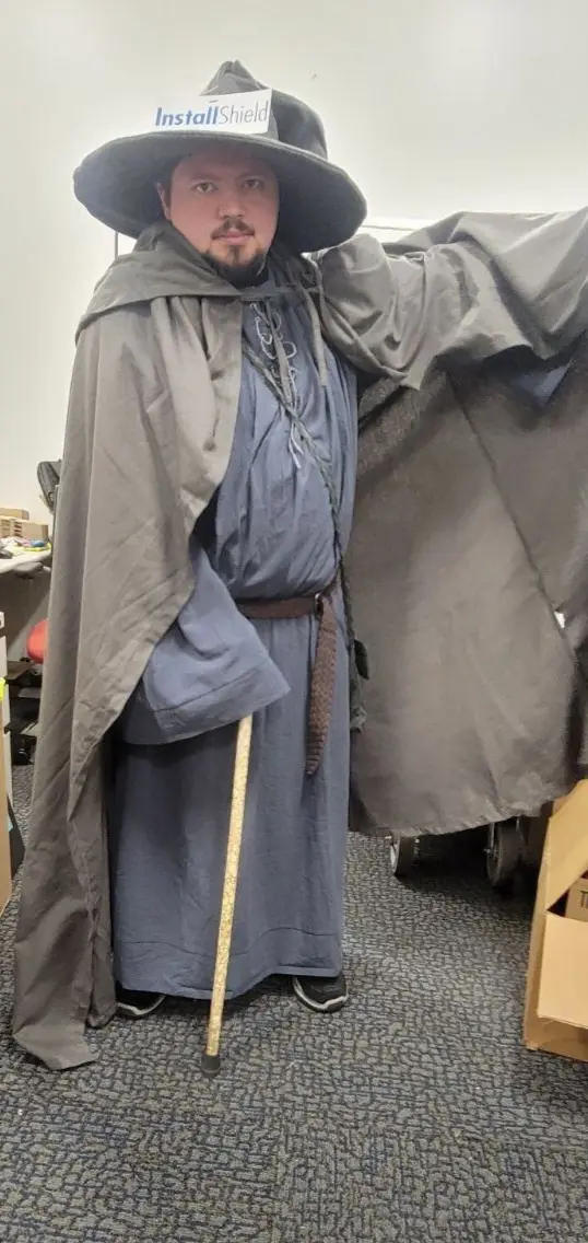 A man dressed as a medieval monk or wizard in a grey robe and wide-brimmed hat stands in an office holding a measuring tape…