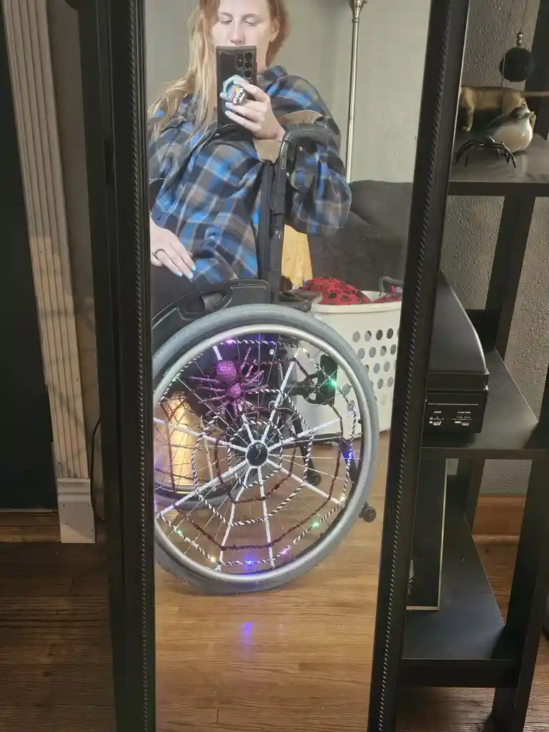 A person in a wheelchair takes a mirror selfie, showing their wheelchair's front wheel decorated with a glittery purple…