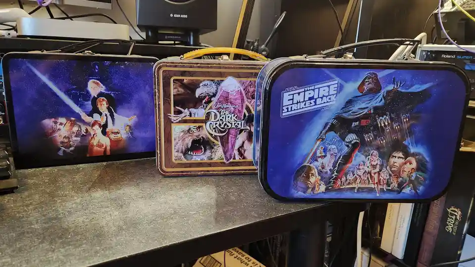Three vintage-style lunchboxes sit side-by-side on a desk, each featuring iconic movie artwork: the left shows a Star Wars…