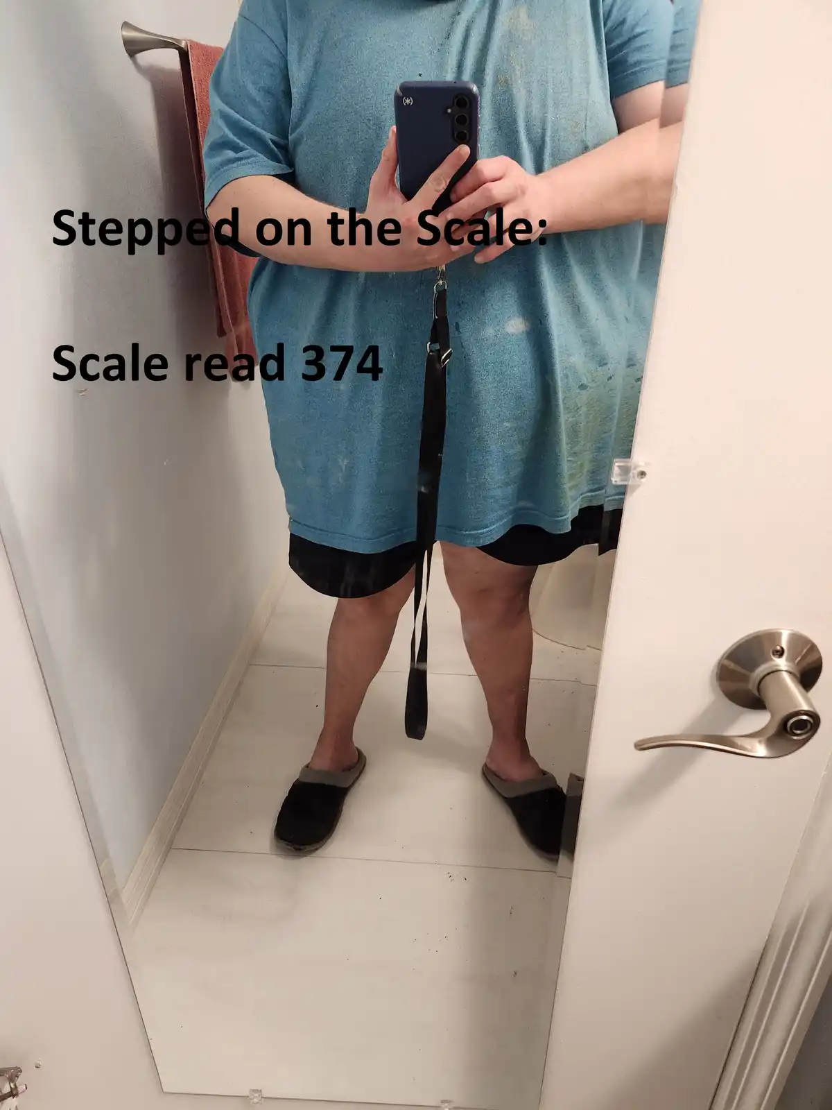 A person stands in a bathroom mirror selfie, wearing a blue t-shirt and black shorts, holding a phone with a leash attached…