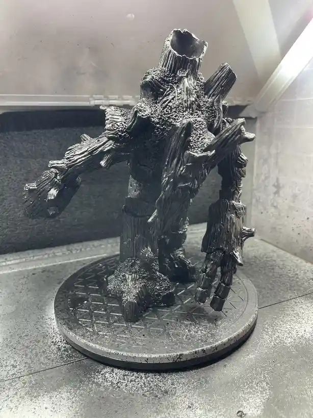 A 3D-printed figurine resembling a humanoid tree, with bark-like skin, branches for limbs, and a hollowed-out head, stands…