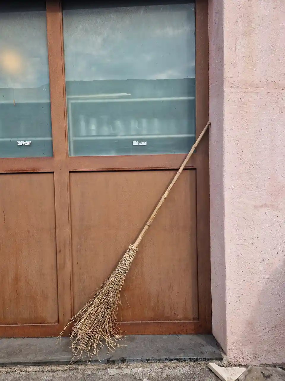 A rustic broom with a bamboo handle leans against a weathered brown metal door with glass panels, resting on a concrete step…