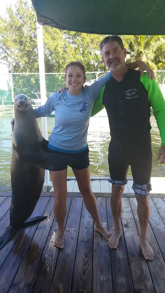 A smiling man and woman stand barefoot on a wooden dock next to a large sea lion that is standing upright and smiling. The…