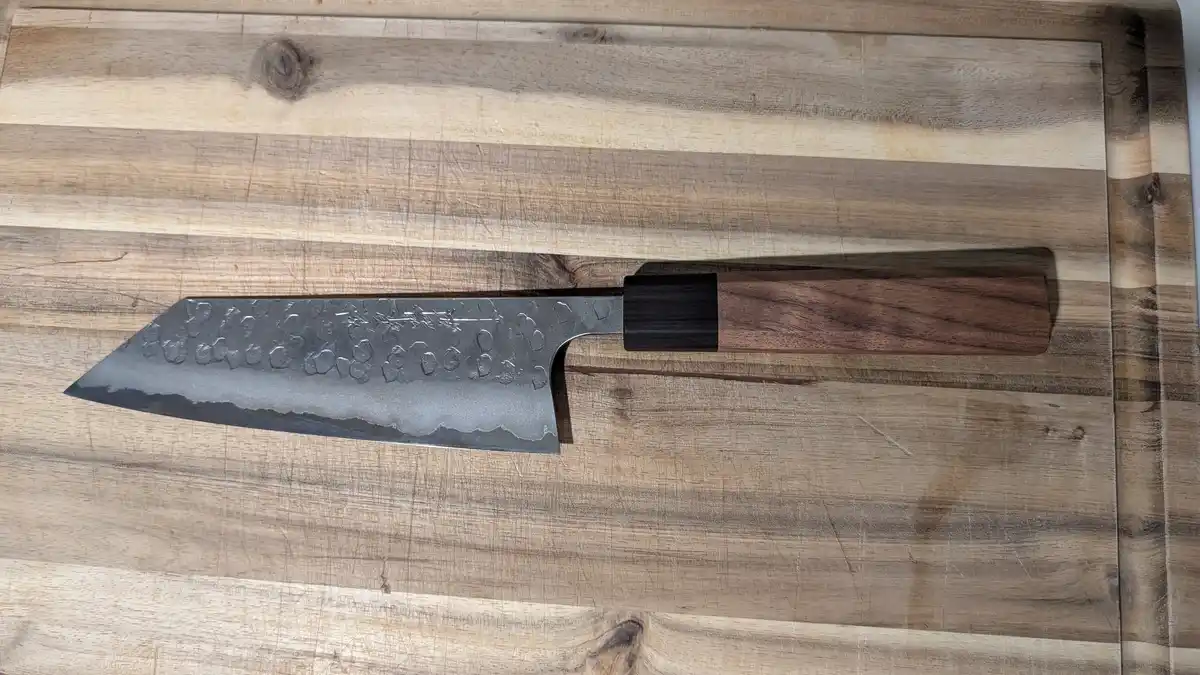 a high-quality chef's knife with a hammered, textured blade and dark wooden handle rests on a well-used wooden cutting…