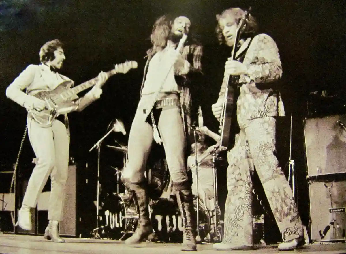 A black-and-white photo of three musicians on stage during a live performance, with one guitarist on the left, a vocalist in…
