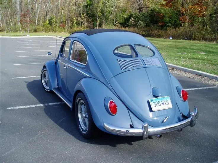A light blue classic Volkswagen Beetle is parked in an asphalt lot with white parking lines, surrounded by grass and trees…