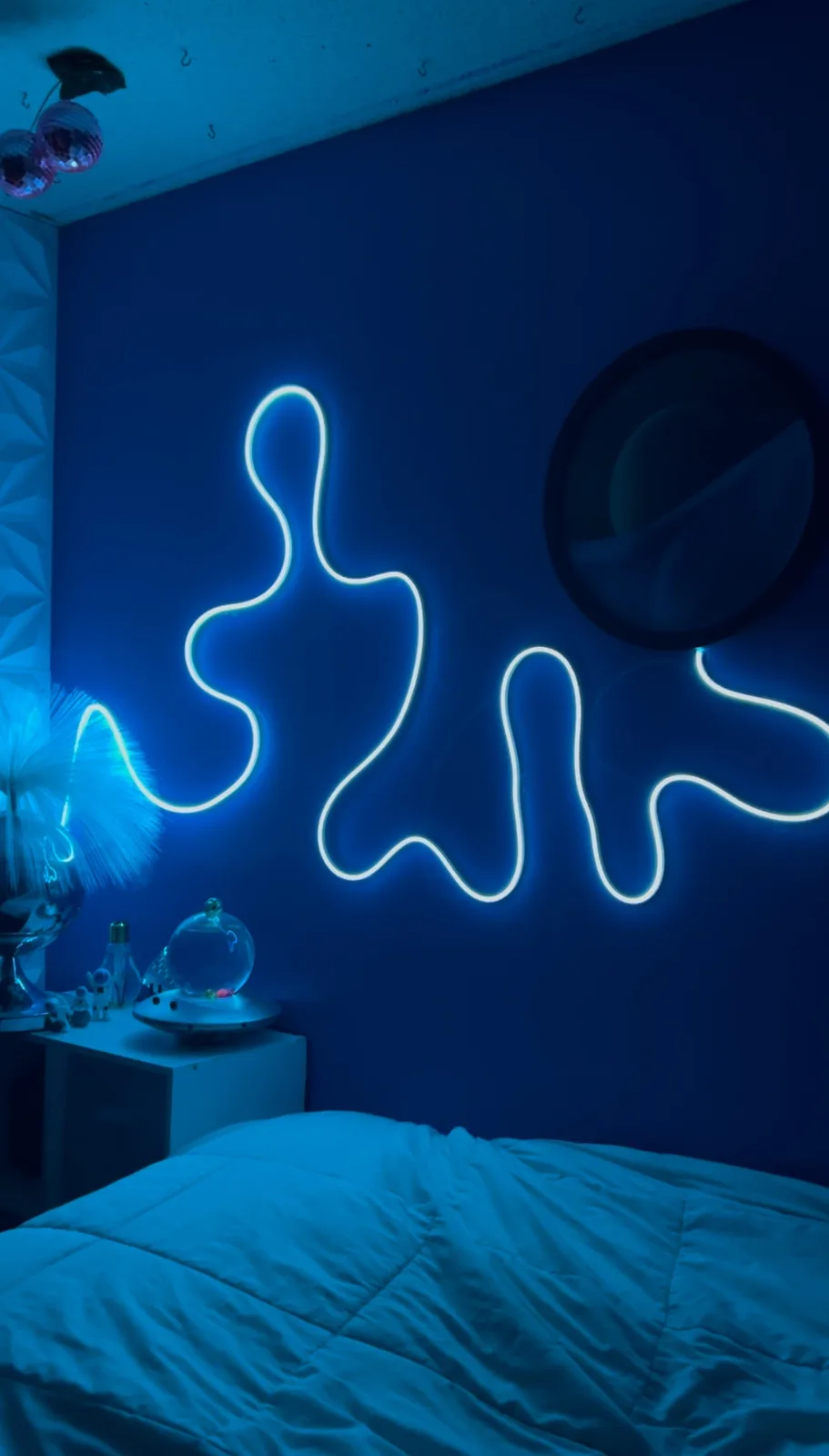 A dimly lit bedroom with blue walls and a glowing white neon light shaped like a wavy, abstract figure resembling a person…