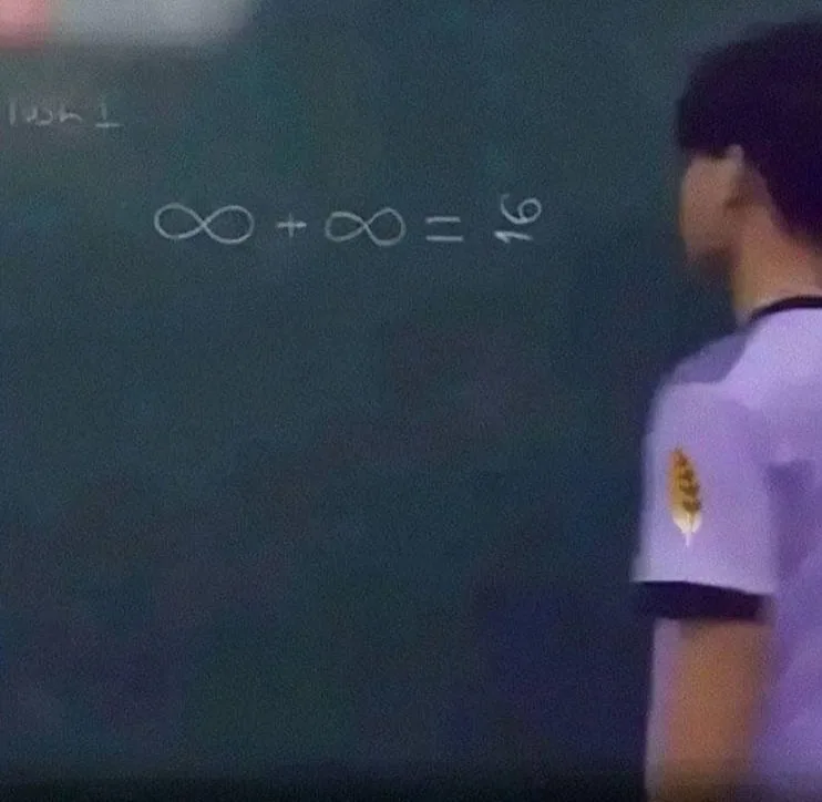 A person in a purple shirt stands beside a dark green chalkboard with white chalk writing. The board displays the equation…