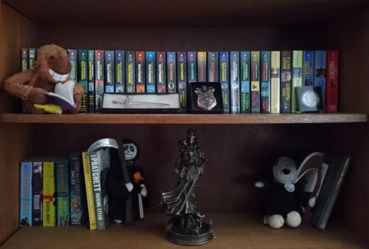 A wooden bookshelf displays numerous Terry Pratchett books, with a brown plush monkey holding a book on the top shelf and…