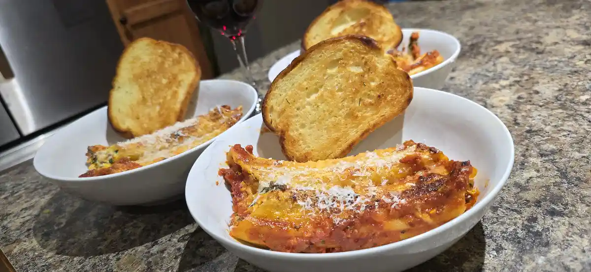 Three white bowls on a granite countertop contain portions of baked lasagna topped with melted cheese and tomato sauce, each…