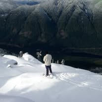 A person stands atop a snowy mountain peak, overlooking a vast valley with other hikers visible in the distance. The image…