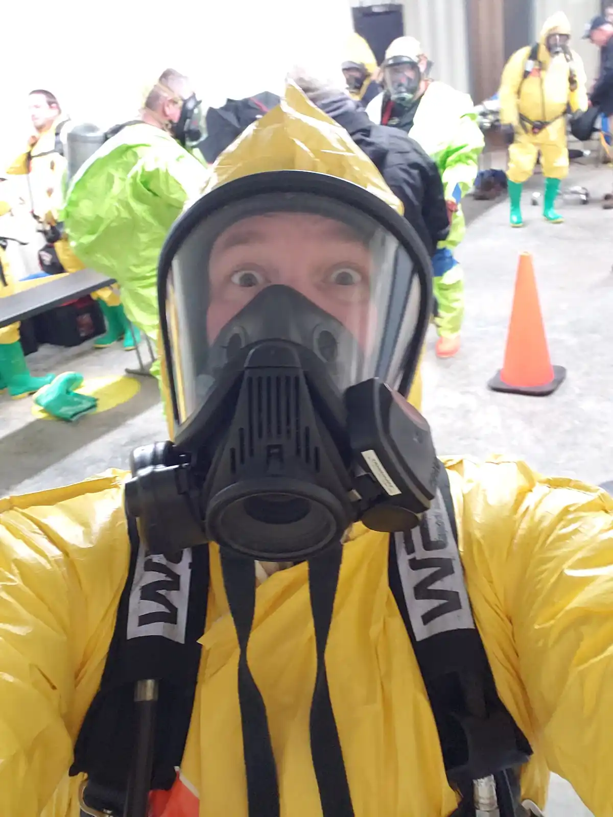 A person in a yellow hazmat suit and black gas mask takes a selfie with wide, startled eyes visible through the mask. Other…