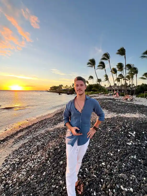 A man stands on a black pebble beach during a vibrant sunset, wearing a blue shirt and white pants, holding a phone. Palm…