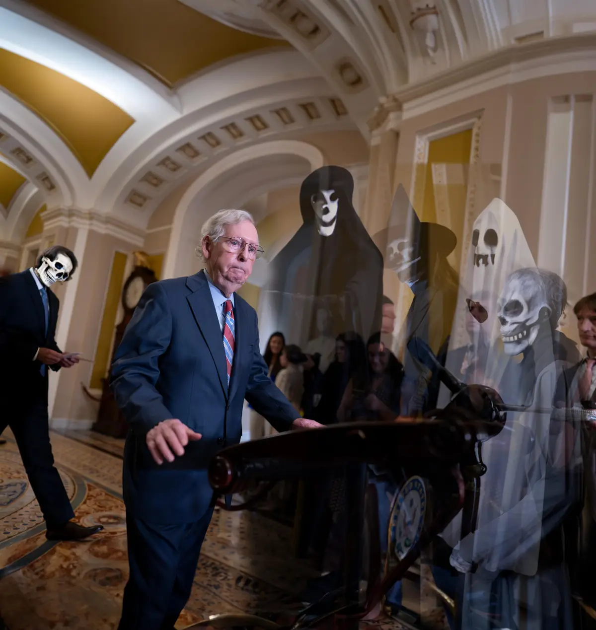 A man in a suit and glasses stands at a podium in a grand, ornate hallway, looking concerned. Superimposed over him are…