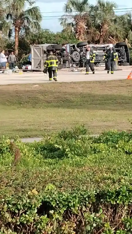 A flipped-over truck lies on its side in a grassy area with palm trees in the background. Three emergency responders in…