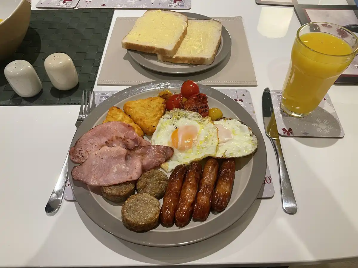 A full English breakfast is laid out on a white table, featuring fried eggs, sausages, ham, hash browns, grilled tomatoes…
