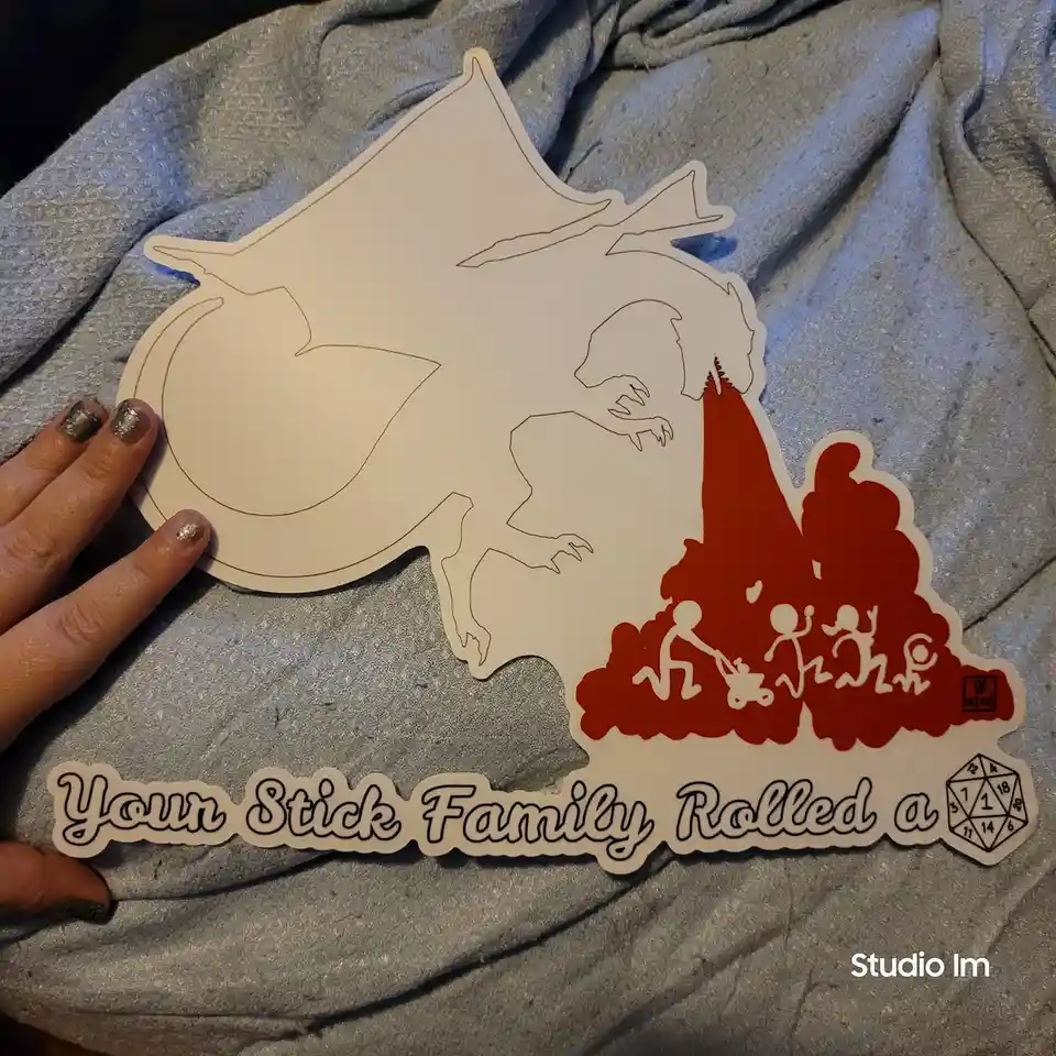 A hand holds a die-cut sticker featuring a large dragon silhouette with a red fire blast engulfing stick-figure family…