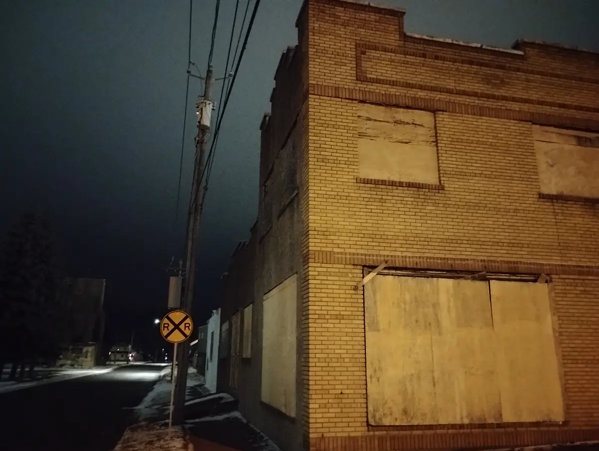 A nighttime street scene shows a yellow railroad crossing sign with 'R R' on it, positioned next to a brick building with…