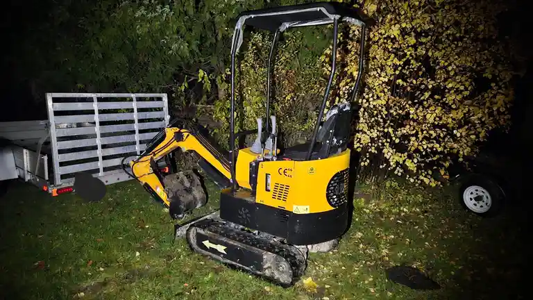 A small yellow mini excavator is parked on grass at night next to a metal trailer and bushes with yellow leaves. The…