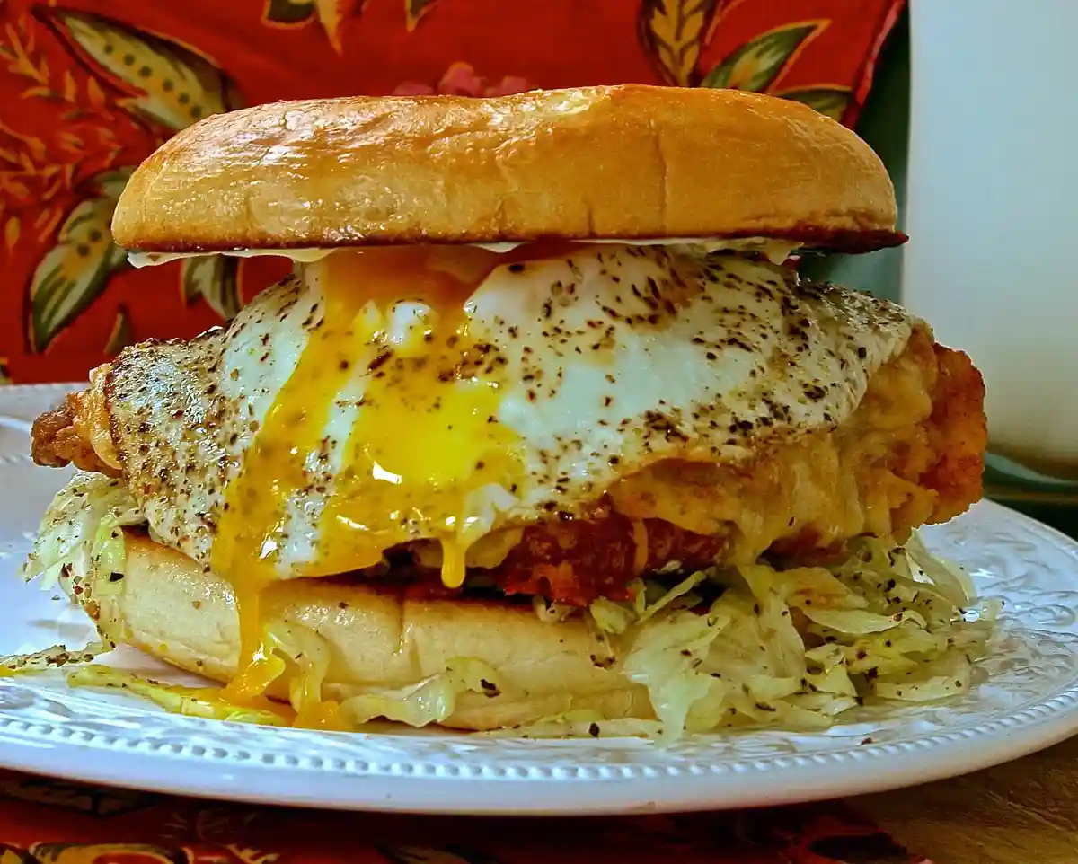 A close-up photo of a sandwich on a white plate, featuring a fried egg with a runny yolk, crispy fried chicken, shredded…