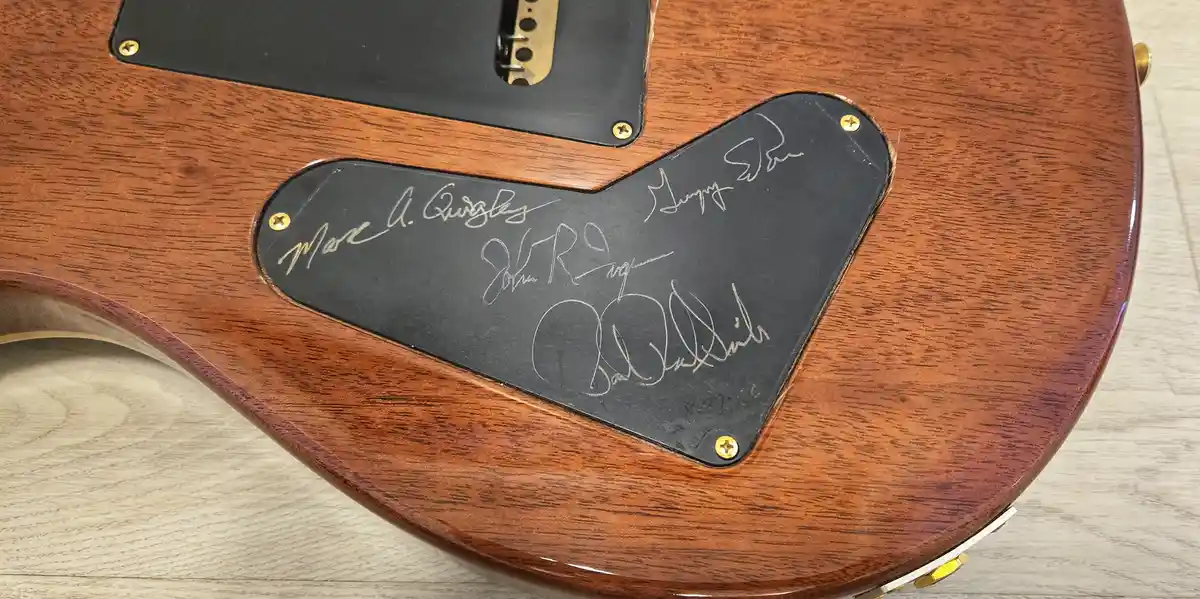 A close-up of a wooden electric guitar's body shows a black pickguard with four handwritten signatures in silver ink. The…