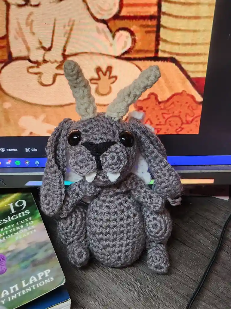 A gray crocheted bunny with long floppy ears, black button eyes, and small white fangs sits on a wooden desk in front of a…