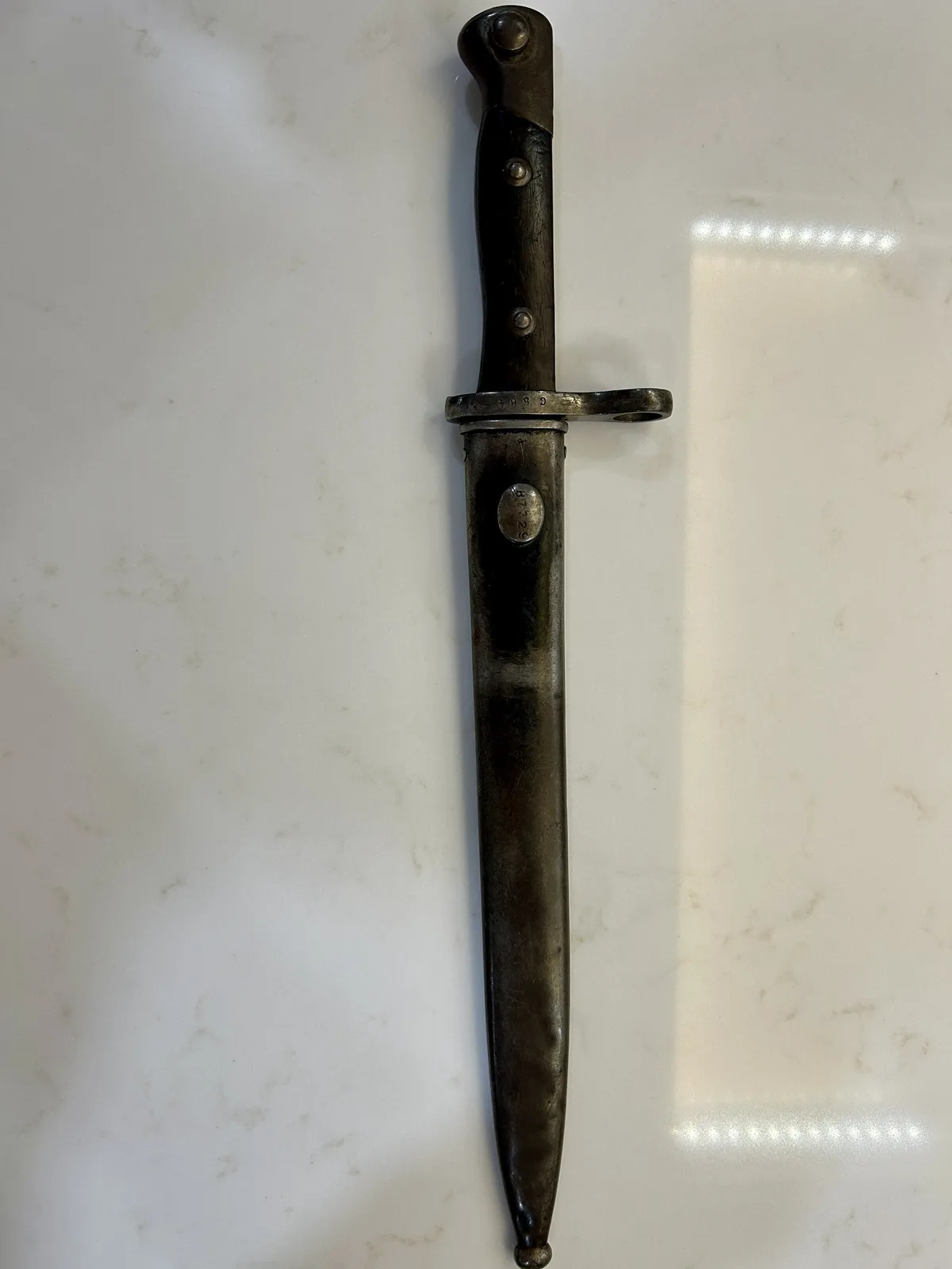A vintage bayonet with a dark wooden handle and metal scabbard rests vertically on a white marble surface. The overlaid text…