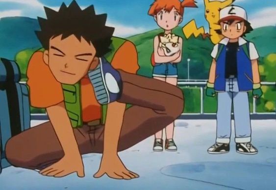 A scene from the anime Pokémon shows Ash Ketchum and Pikachu standing behind a boy in an orange shirt who is sitting on the…