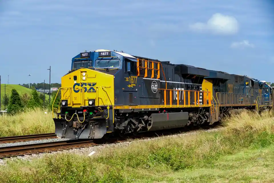 A CSX freight train locomotive, numbered 1877, is shown on tracks with grassy fields and a blue sky in the background. The…