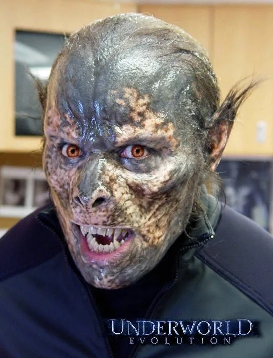 A person wearing a detailed, grotesque werewolf or vampire-like makeup with textured gray and brown skin, pointed ears, and…