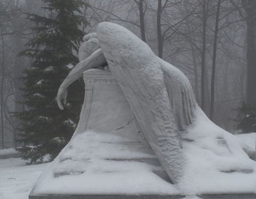 A snow-covered stone statue of a weeping angel or figure, head bowed and arms draped over its chest, set in a snowy, wooded…