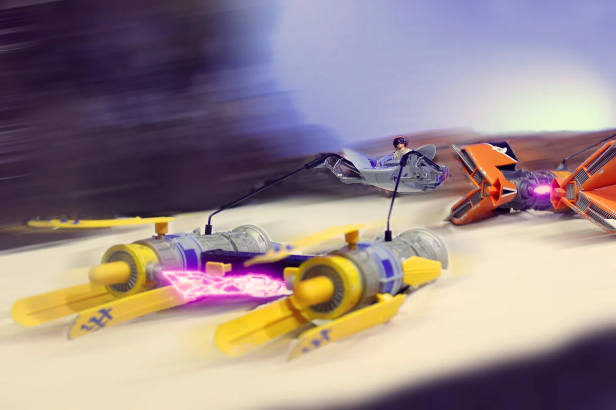 A dynamic, motion-blurred photo shows three toy-like futuristic racing vehicles speeding across a desert-like surface. The…