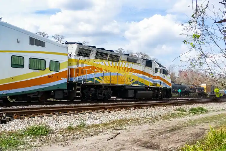 A colorful train with a yellow, orange, and green sunburst design on its locomotive is shown on tracks under a partly cloudy…