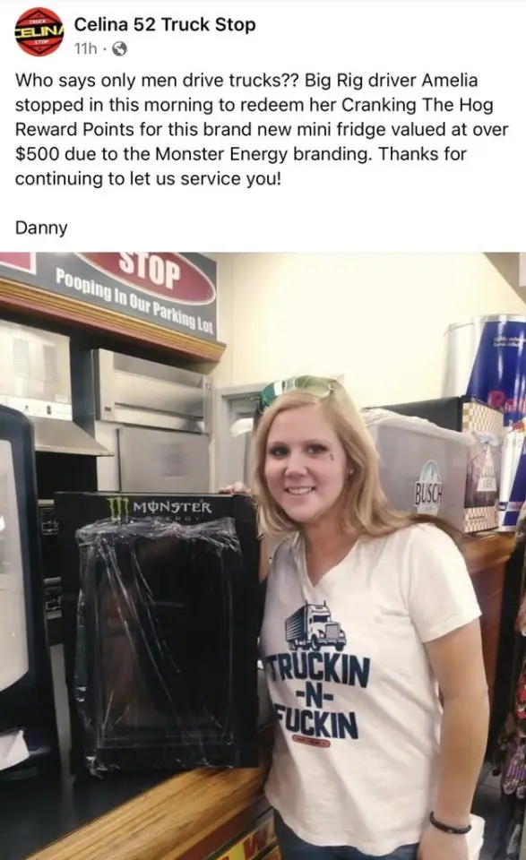A blonde woman in a white t-shirt with a truck graphic stands smiling beside a wrapped Monster Energy-branded mini fridge at…