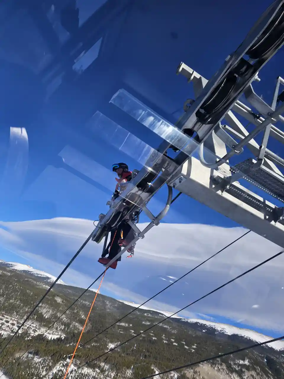 A person wearing a helmet and safety gear is suspended on a ski lift mechanism high above a snowy mountain landscape under a…