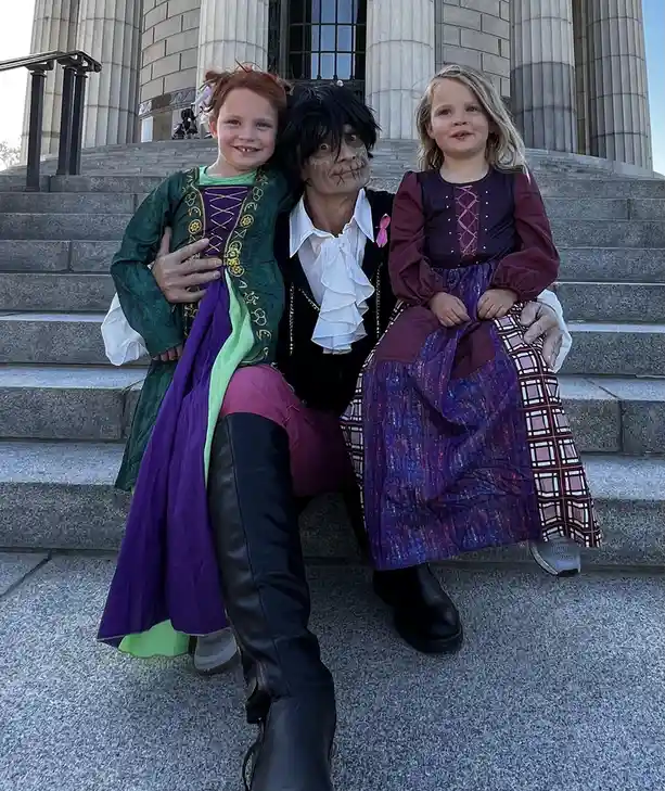 Three people pose on stone steps in front of a classical building: a man in a skeletal makeup costume with a white ruffled…