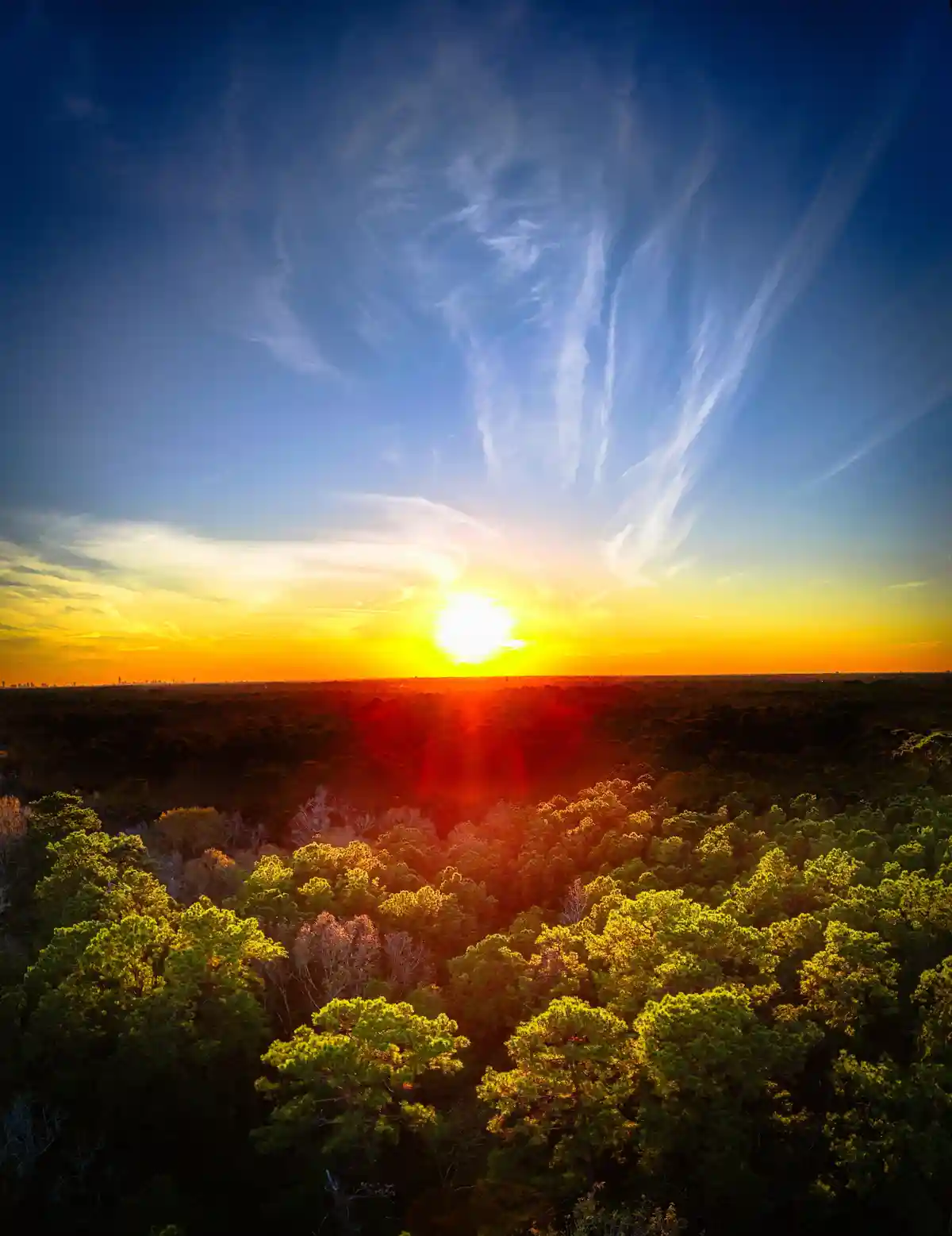 Aerial view of a vibrant sunset over a dense forest, with the sun low on the horizon casting golden light and red lens…